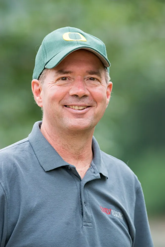 A headshot of John Heldt in a gray polo and green had with the Oregon O on the front.