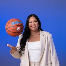 Malena Udani throws a basketball in the air while wearing a tan suit in front of a royal blue background.