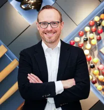 A person smiling while crossing their arms in front of a modern decorated wall. 