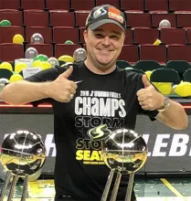 Person giving two thumbs up while wearing a championship T‑shirt and cap, standing behind two trophy awards in an arena decorated with team-colored balloons.