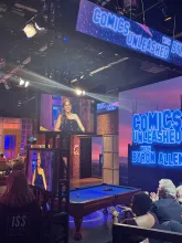 Screens on a TV show set say COMICS UNLEASHED WITH BYRON ALLEN. A white woman in a black dress with long brown hair is displayed on monitoring screens while she smiles.