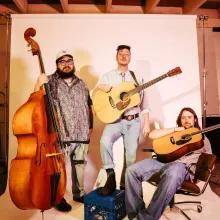One man holds a standing bass and two men hold guitars in front of a white photo background in orange light.