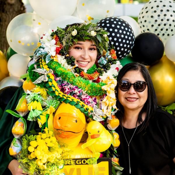 A young woman covered in leis stands next to her mom in front of balloons.