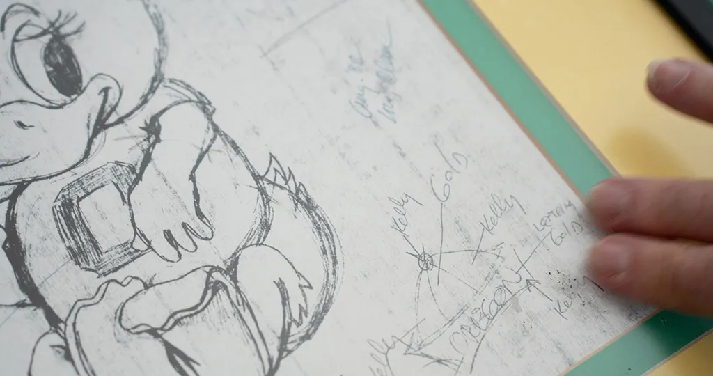 Hand pointing to a sketch of a cartoon duck in an 'O' jersey with handwritten notes.