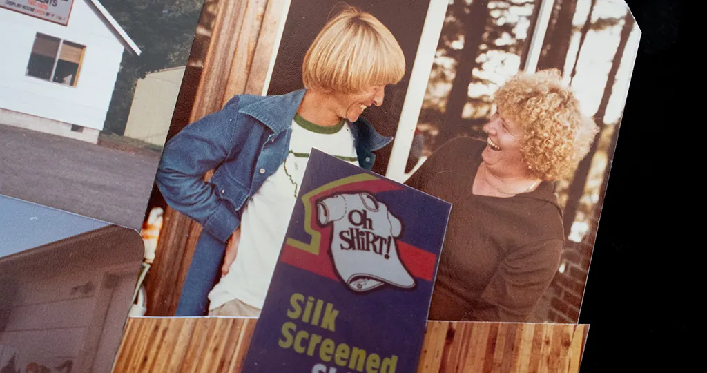 Two people behind an 'Oh Shirt!' silk-screened shirt advertisement.