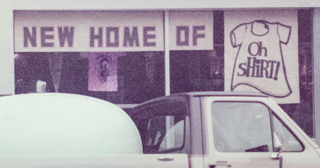 A truck driving by a print shop with a banner that reads, "New Home of Oh Shirt!"