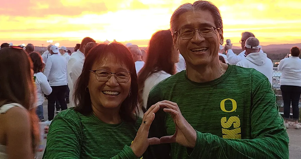 June and Sam Liu make an O with their hands together while Penn State fans in white roam around behind them. The sunset is a brilliant shining orange behind them.