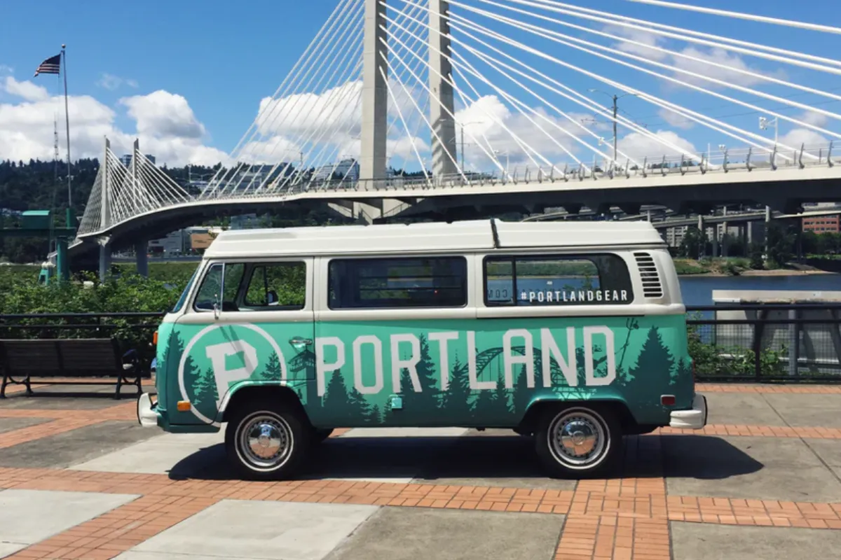 Green and white Portland-themed van parked near Tilikum Crossing bridge under a partly cloudy sky.