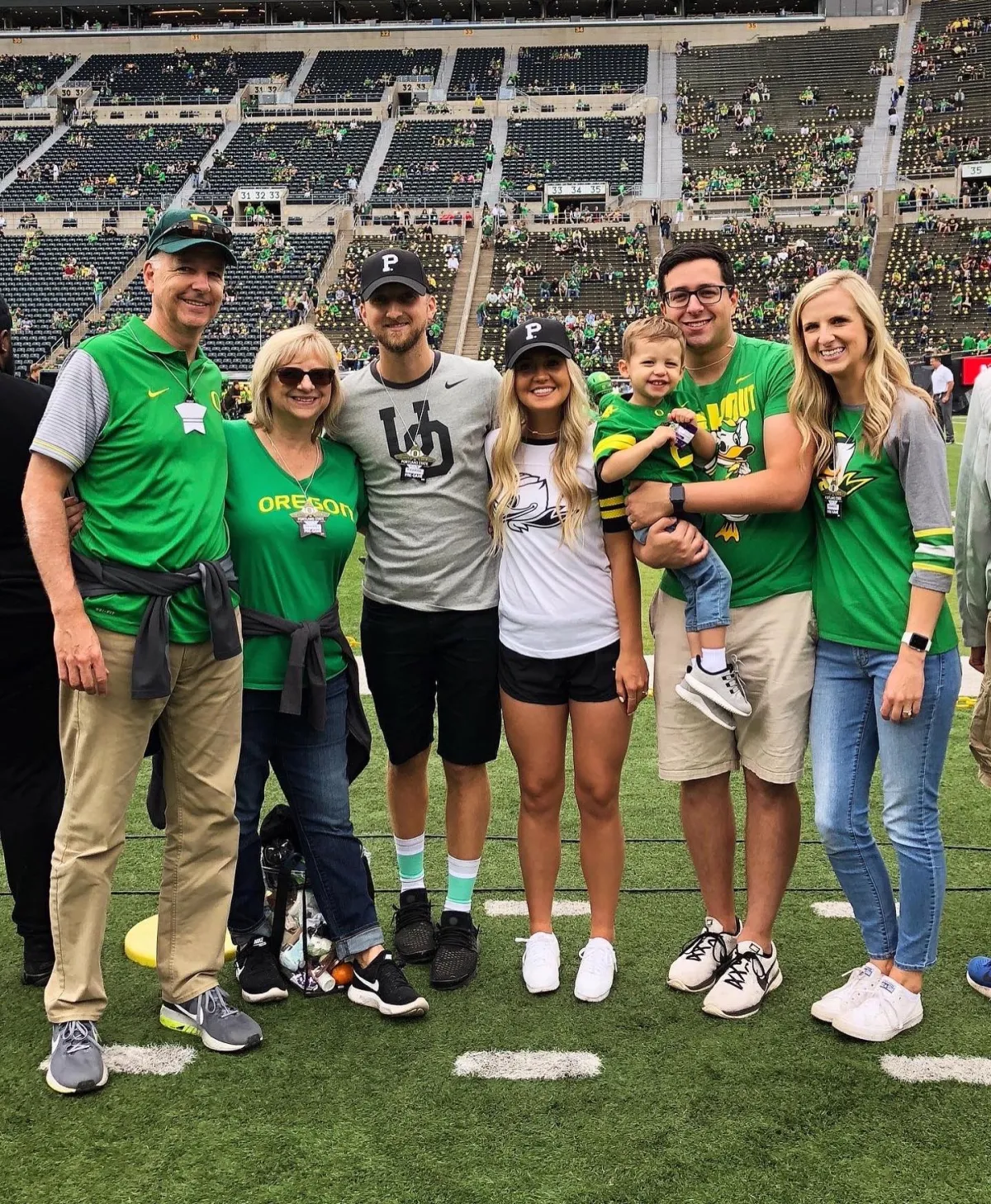 Two adults and two children in Oregon Ducks gear on a football field with a stadium crowd in the background.