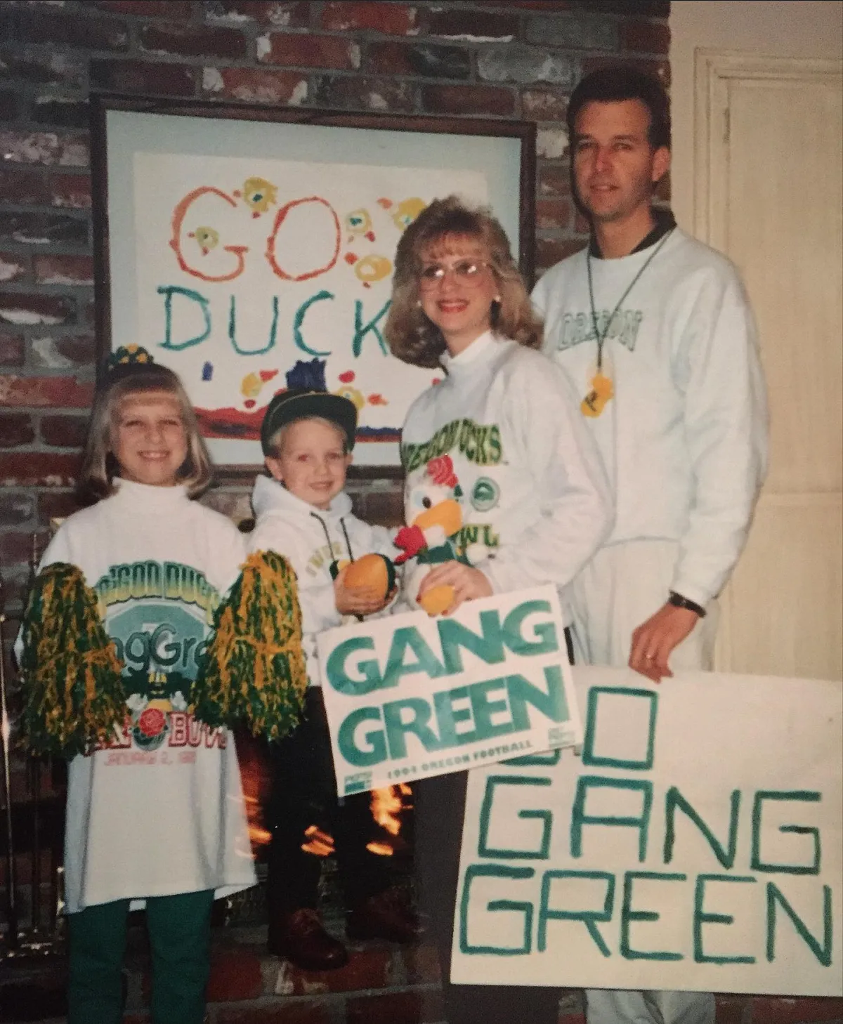 Group of four people in Oregon Ducks gear holding 'Gang Green' signs in front of a 'Go Ducks' wall.