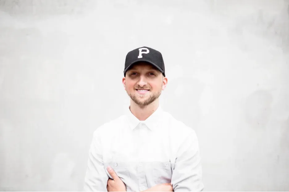 Person in a white shirt and black 'P' cap standing with arms crossed against a plain background.