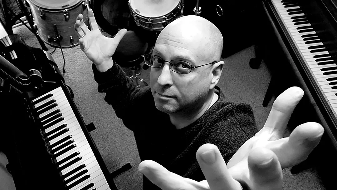 Person standing in a music room with keyboards and drums, arms raised, black and white image.