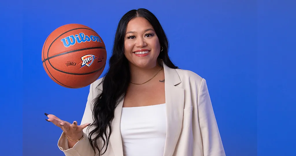Malena Udani throws a basketball in the air while wearing a tan suit in front of a royal blue background.