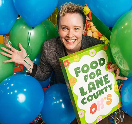 A white female standing in a frame of balloons holding a box that says "Food for Lane County."