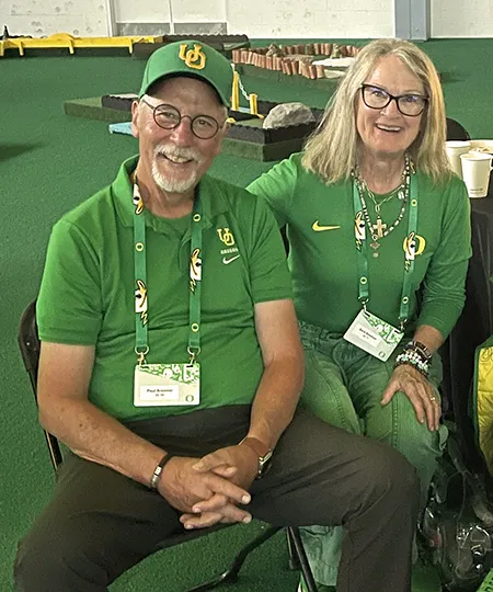 Two people seated indoors wearing matching green University of Oregon apparel and event lanyards.
