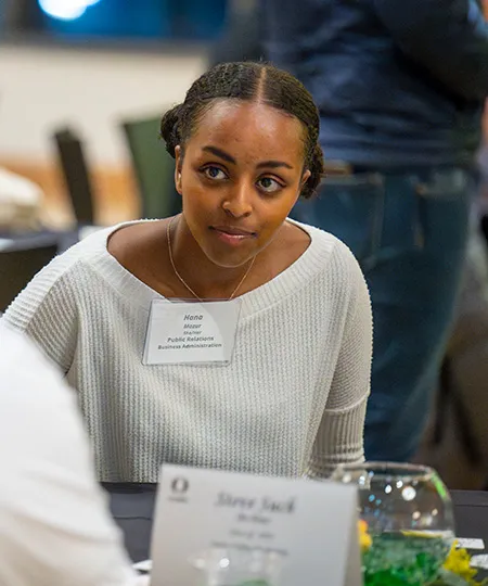 A student seated at a table wearing a white sweater and a name badge during an indoor event.