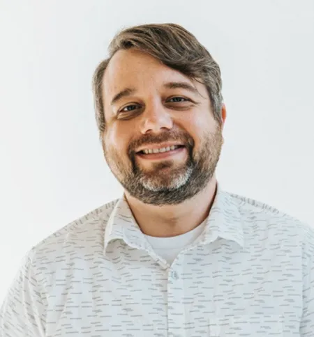 A white male with curvy combed hair and beard smiling wearing a white shirt. 