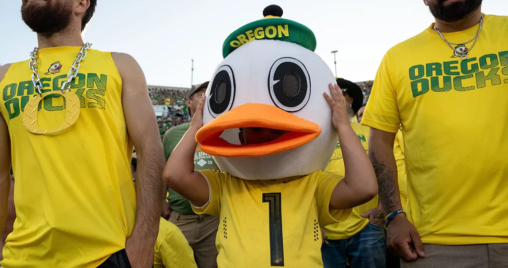 A child in a yellow Oregon jersey wears an Oregon Duck head while adults on both sides of them wear yellow shirts that say Oregon Ducks.