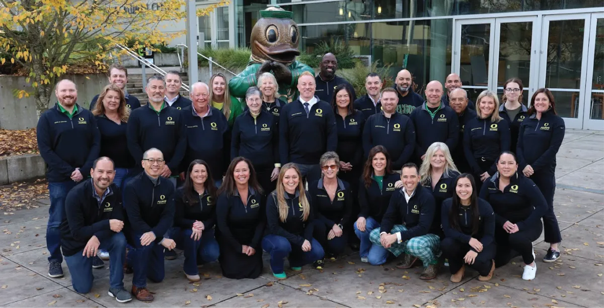 A group picture of the University of Oregon Alumni Association Board Members, all wearing a black long-sleeve quarter zip, posing in front of a Duck statue. 