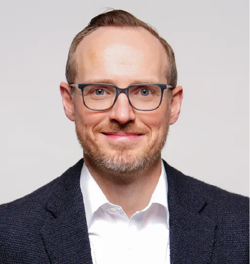 A headshot of a person wearing glasses and a blue pullover in front of a white background. 