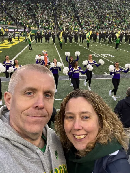 James and Emma grin at the camera in a selfie close to the field in front of a group of cheerleaders in Autzen Stadium.
