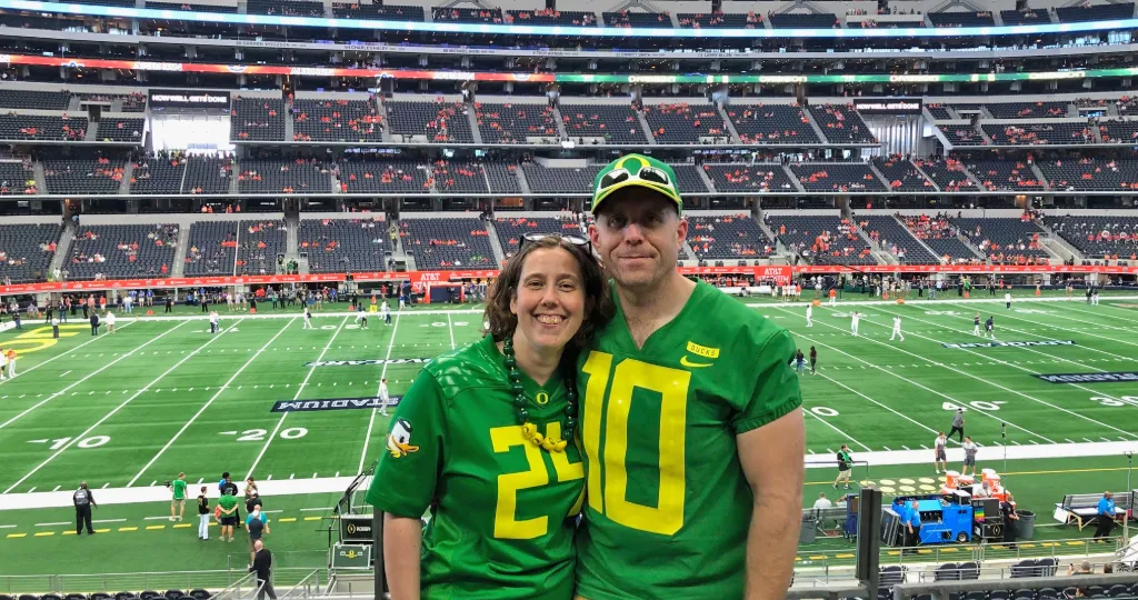 James and Emma Hathaway pose close together wearing bright green Oregon Ducks football jerseys with the inside of a football stadium behind them.