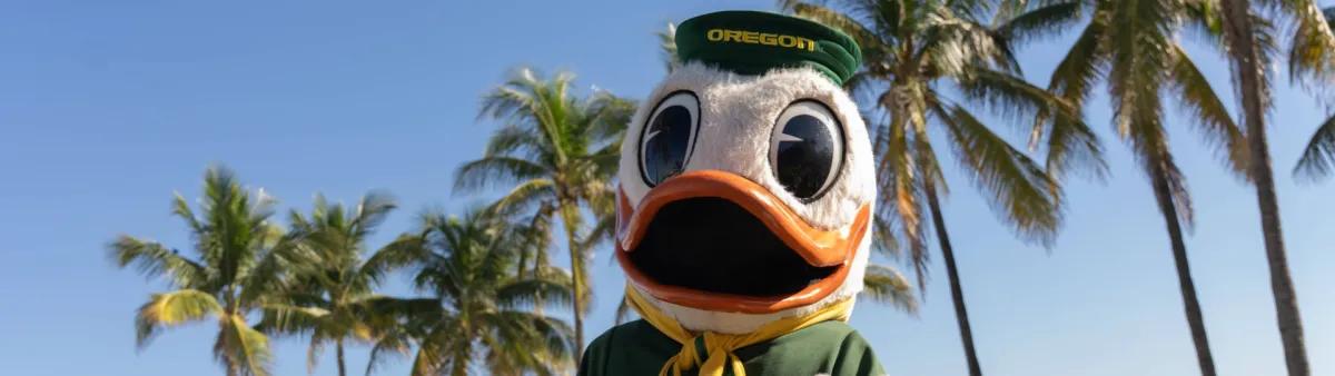 The Oregon Duck poses in front of a blue sky and palm trees.