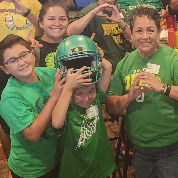 Stacey Moniz poses with two young boys, one of whom is putting on a green Ducks football helmet.