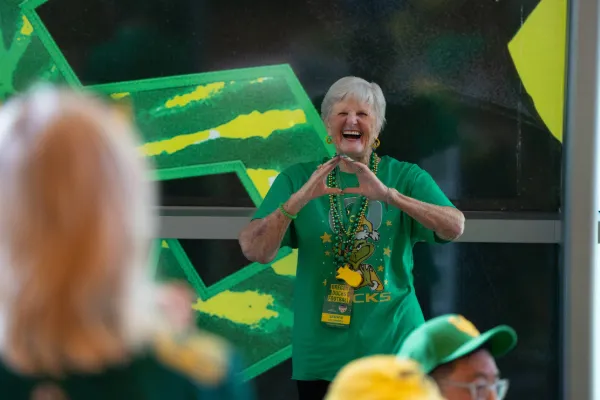 An older woman with short gray hair wears a green Oregon shirt, and green and yellow beads around her neck while making an O with her hands and smiling at the camera.