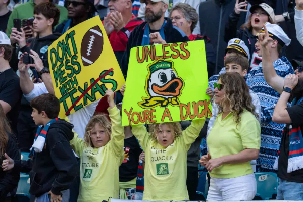 Two children in yellow long sleeve shirts hold signs above their heads. The one on the left says Don't Mess with Texas with a red line crossing out Texas, and the right sign says Unless You're a Duck, with the Oregon Ducks logo in the center.