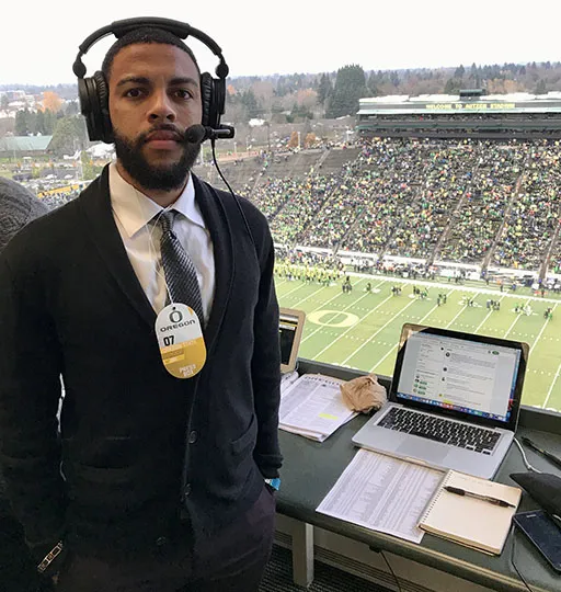 A person standing in a reporting booth during a football game while wearing a headset and suit. 