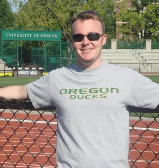 erson leaning against a chain-link fence on a track field, wearing an ‘Oregon Ducks’ T‑shirt with stadium seating and campus signage in the background.