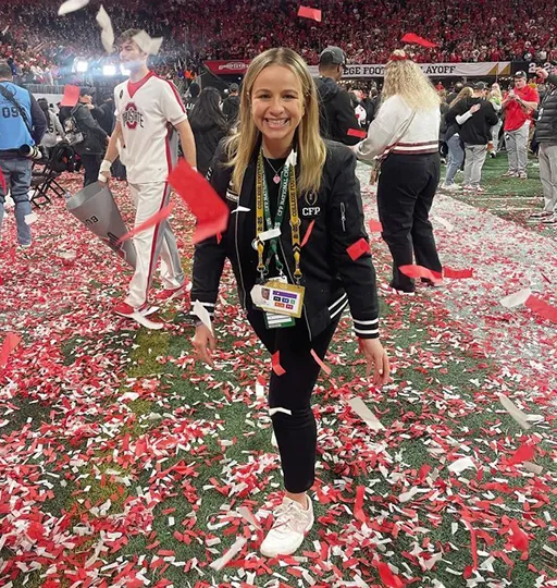 Person walking across a football field covered in red and white confetti after a championship game, wearing event credentials, with a celebrating crowd in the background.sense of 