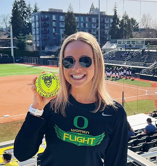 Person holding a yellow softball at an outdoor stadium, wearing a black ‘Oregon Women Flight’ shirt.