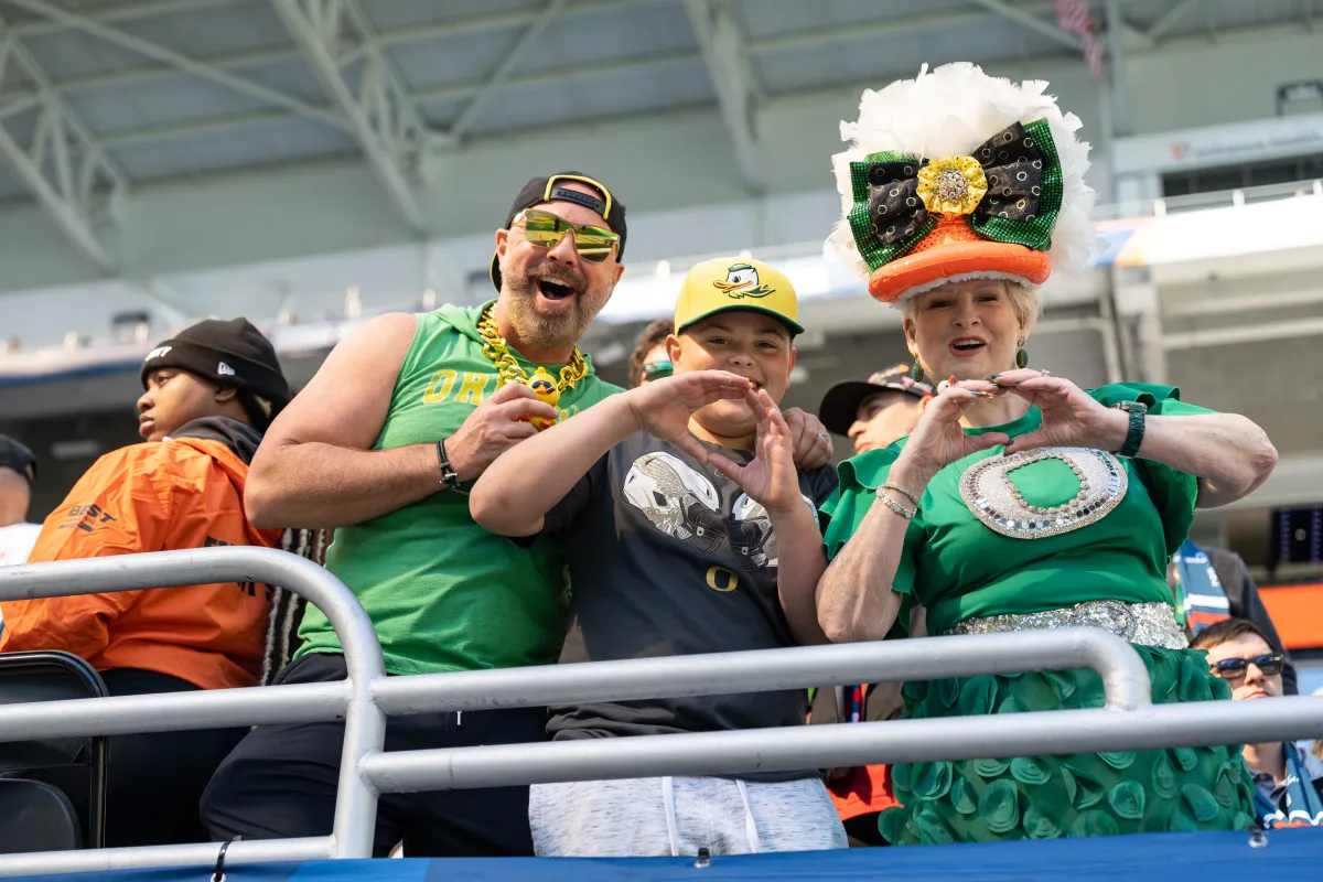 Fans in green and yellow Oregon Ducks gear pose together in the stadium stands, making hand signs and wearing festive team outfits.