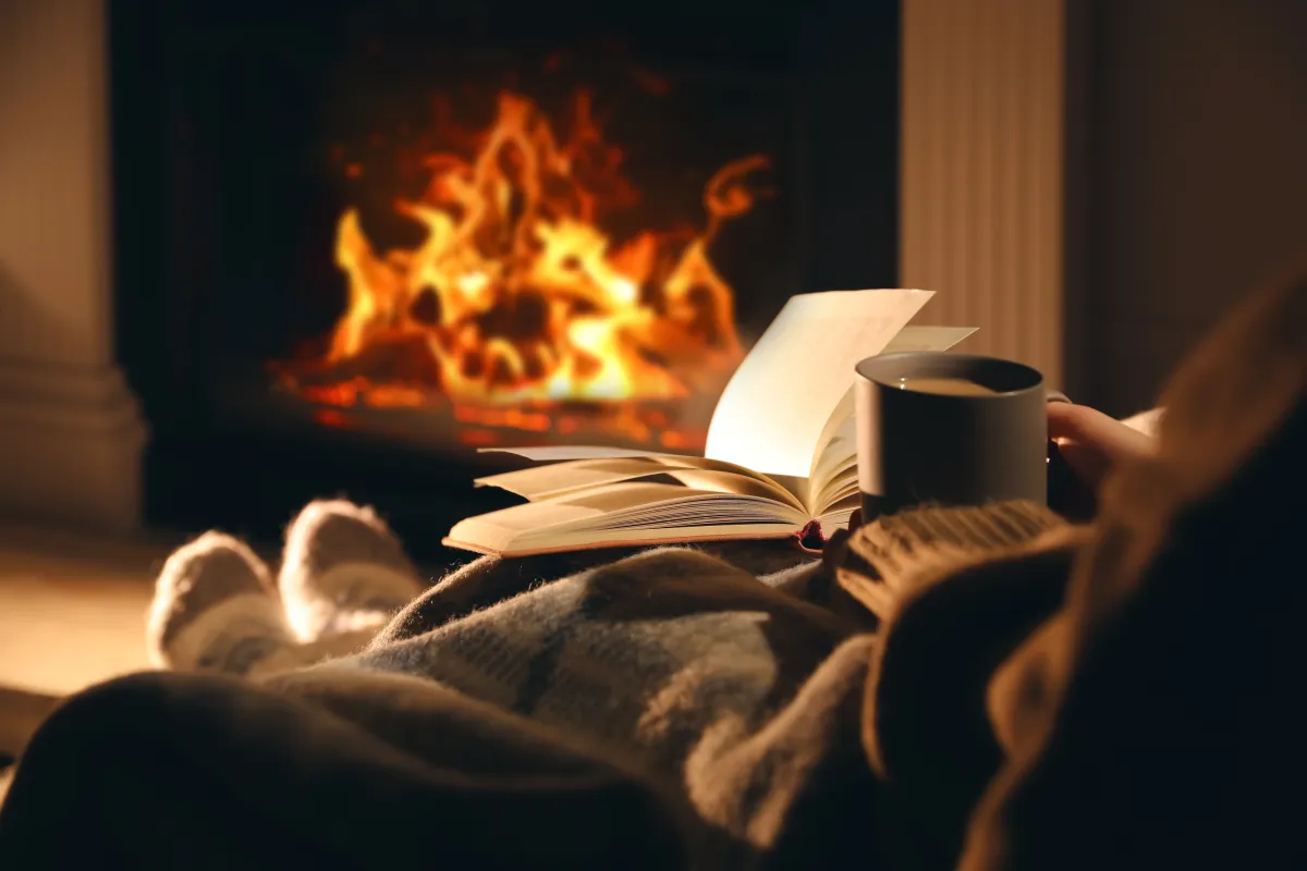 A cozy person sitting down reading a book and drinking a warm beverage in front of a fireplace. 