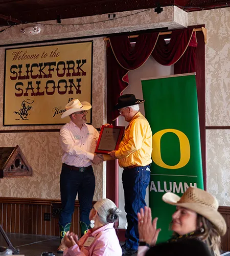 On a stage in front of a green banner with a yellow O that says Alumni, UOAA Executive Director Raphe Beck hands an award plaque to Senator Bill Hansell while shaking hands. They are both wearing cowboy hats, button up shirts, and jeans.