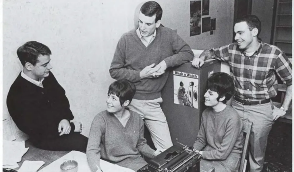 A black and white film photo of three young men and two young women in mid-1960s styling sit and stand at a desk in an office, smiling while talking.