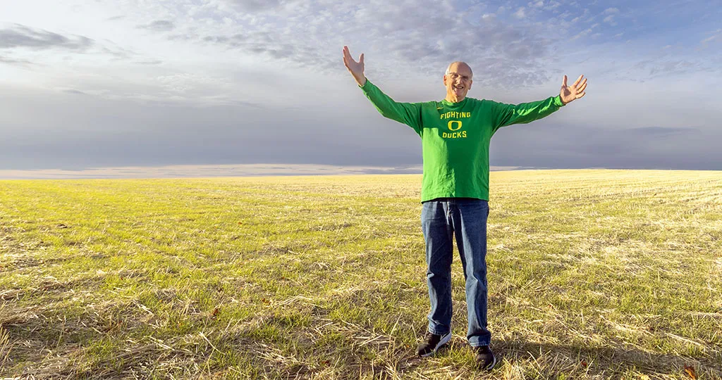 An older white gentleman, Senator Bill Hansell, spreads his harms wide in a yellow grass field, the blue sky with clouds spreading behind him. He is in a bright green shirt that says Fighting Ducks with a yellow O in the middle.