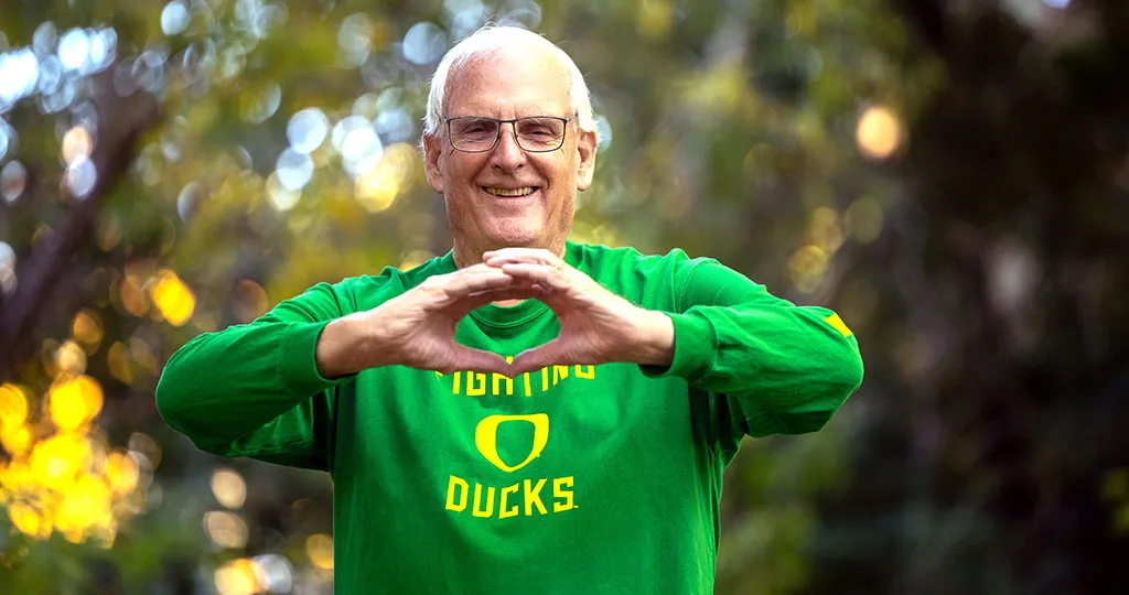 An older white gentleman, Senator Bill Hansell, makes an O with his hands, wearing a bright green long sleeve shirt that says Oregon Ducks in yellow. Blurred trees are behind him.