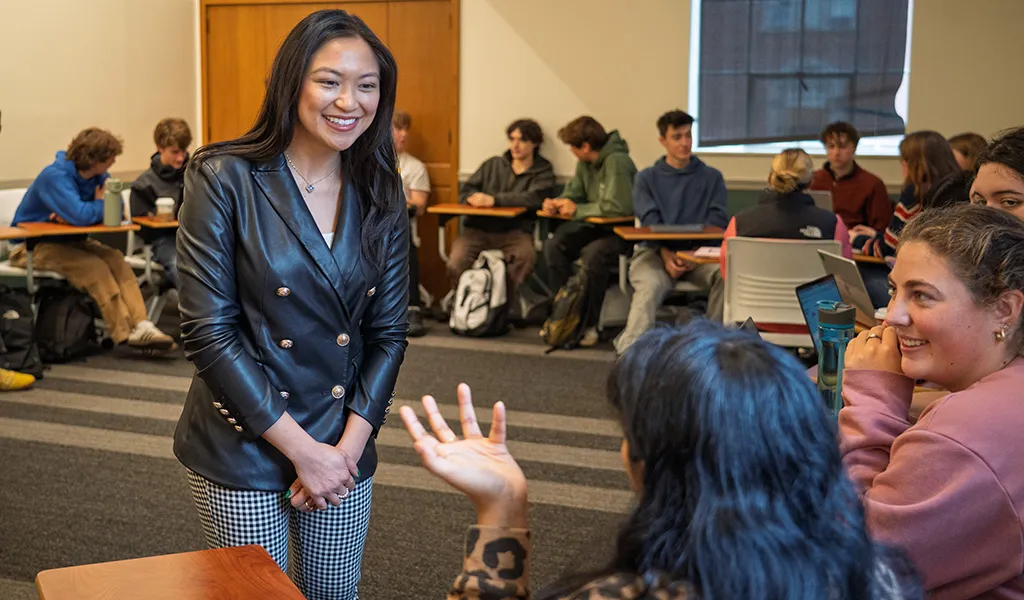 Lorie Acio stands in the middle of a circle of desks with UO students, a student asking her a question while she smiles, engaged.