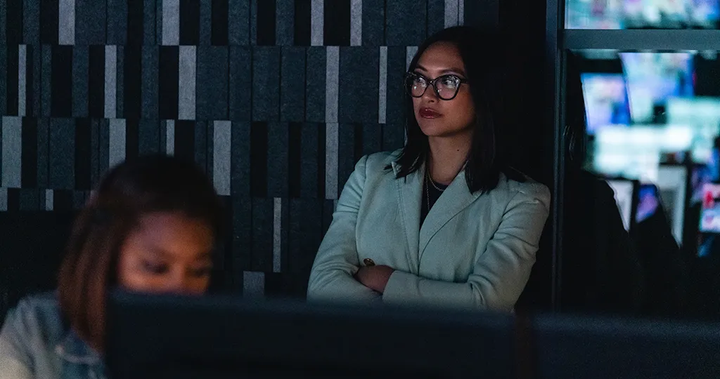 Lorie Acio stands with her arms crossed at the back of an NBC newsroom, the reflections of production screens in the windows behind her while she looks on.