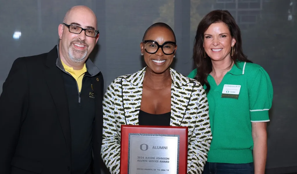 A bald smiling middle-aged white man stands on the left, smiling Black woman with a shaved head and black thick rimmed glasses stands in the center holding a plaque that says "2024 Jeanne Johnson Alumni Service Award Ericka Warren, BA ’92,MBA ’19, and a white woman with brown hair in a green shirt stands on the right, smiling.