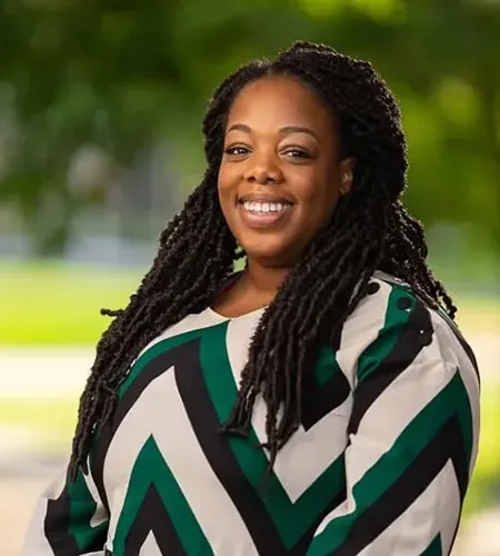 A Black woman with long locs down her shoulders poses for a headshot, wearing a black, green, and white chevron blouse in front of a blurred natural background.