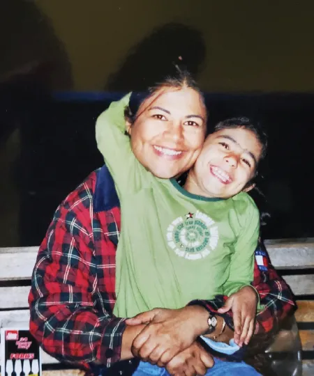 A mom wearing a flannel while sitting on a bench with her daughter sitting on her lap wearing a green long sleeve and her arm wrapped around her moms neck. 