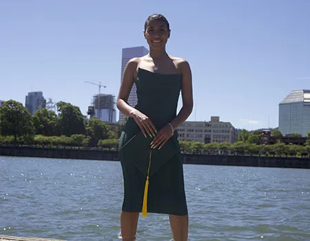 Nia Pennington stands on the waterfront in Portland with the skyline behind her. She is wearing a green dress and holding a graduation cap.