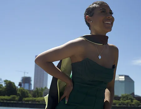 Nia Pennington smiles wearing a green dress and her master's degree cape in front of the Portland skyline on a waterfront.