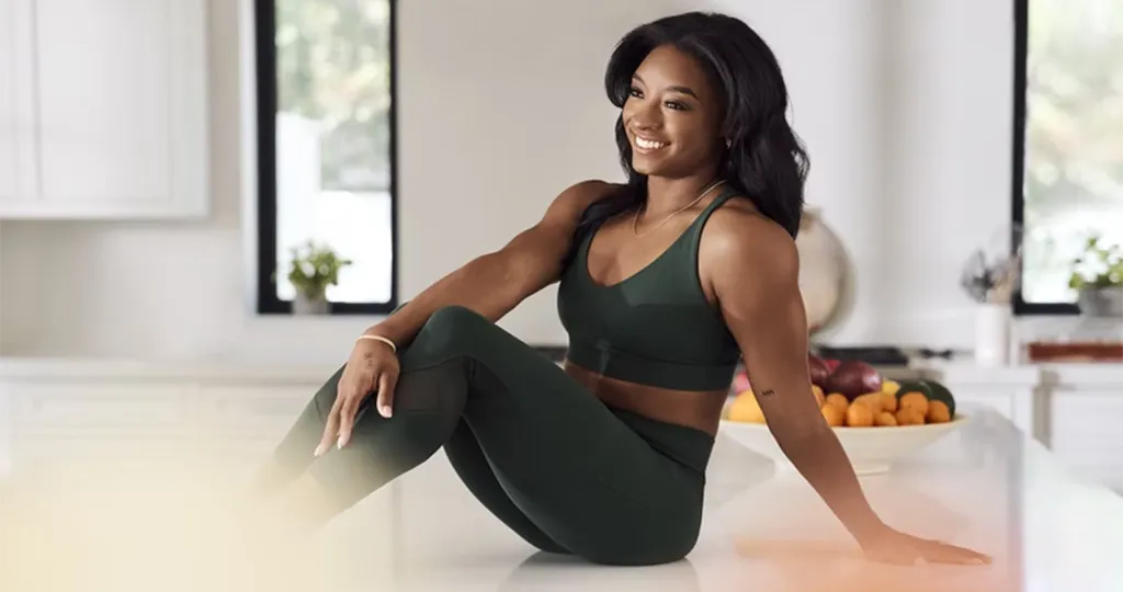 Simone Biles sits on a white granite base in dark green leggings and a matching sports bra that were designed by Nia Pennington for Athleta.