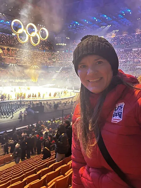Ashley Kirsininkas wears a red coat and a black beanie while the Olympic Rings hang from the ceiling of an Olympic Stadium in Milan, Italy during the Opening Ceremony.