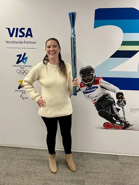 Ashley Kirsininkas holds a chrome blue Olympic Torch in front of the Visa sponsorship wall in her office at the Milano-Cortina Olympics.